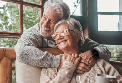 Two elderly individuals sharing a warm embrace while seated, with a cup on the table and a window showing greenery in the background.